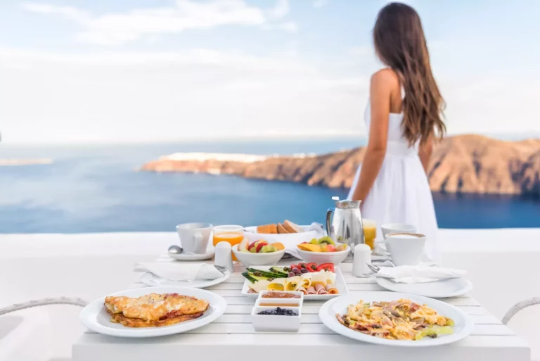 Ontbijttafel en luxe reisvrouw op santorini. Goed uitgebalanceerde perfecte ontbijttafel geserveerd in resort. Vrouwelijke toerist kijkt naar prachtig uitzicht op zee en caldera genietend van haar vakantie.