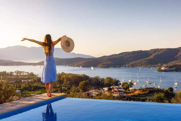 En glad feriekvinde i kjole står ved swimmingpoolen og nyder sommersolnedgangen bag Middelhavet.